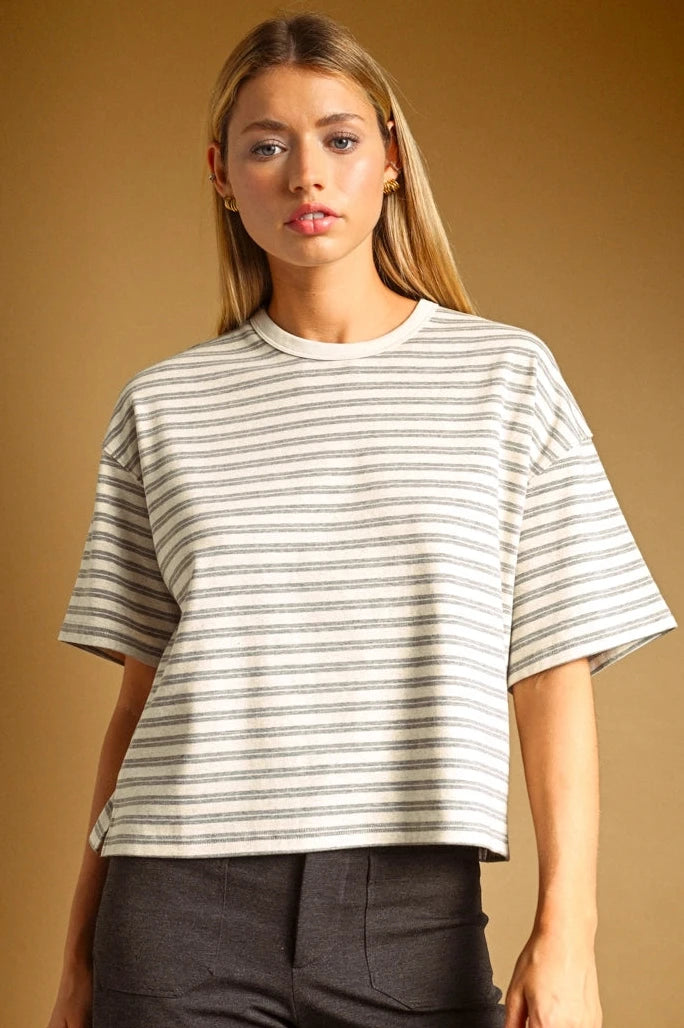 A woman modeling a short-sleeve crewneck t-shirt with thin horizontal gray stripes on an off-white base, paired with dark gray pants, against a solid warm brown studio background.