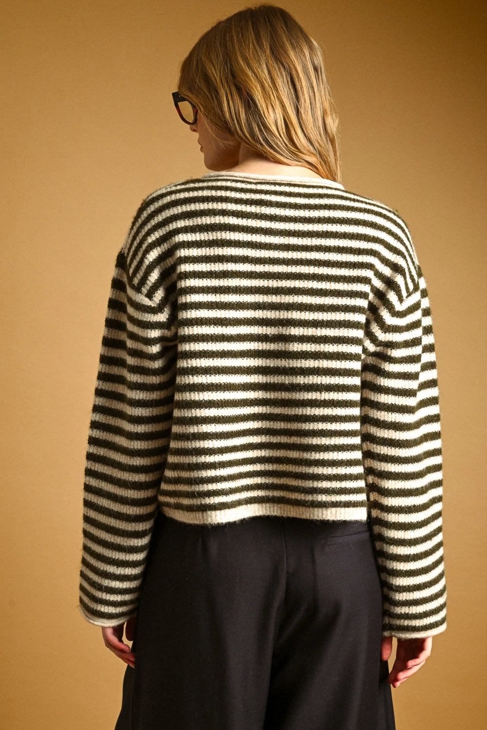 A woman wearing thick black-framed glasses and an oversized black and cream striped knit button-front top, posed against a warm tan studio background.