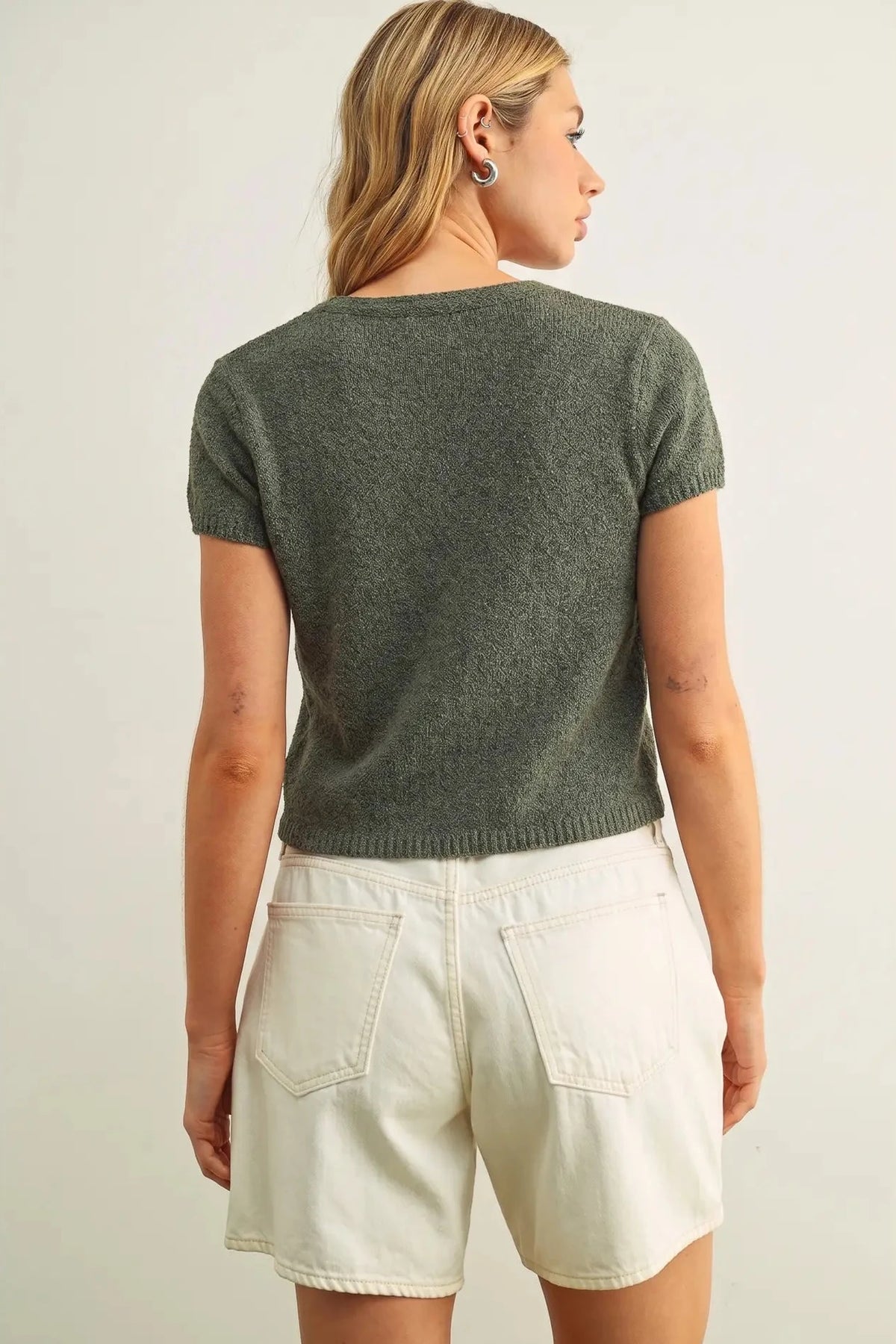 Woman wearing a green knit top and white shorts on a plain background