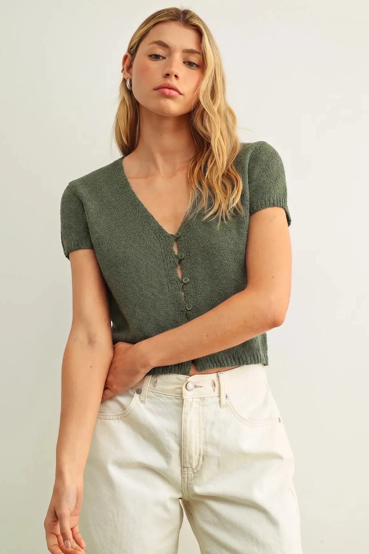 Woman wearing a green knit top and light-colored pants against a plain background