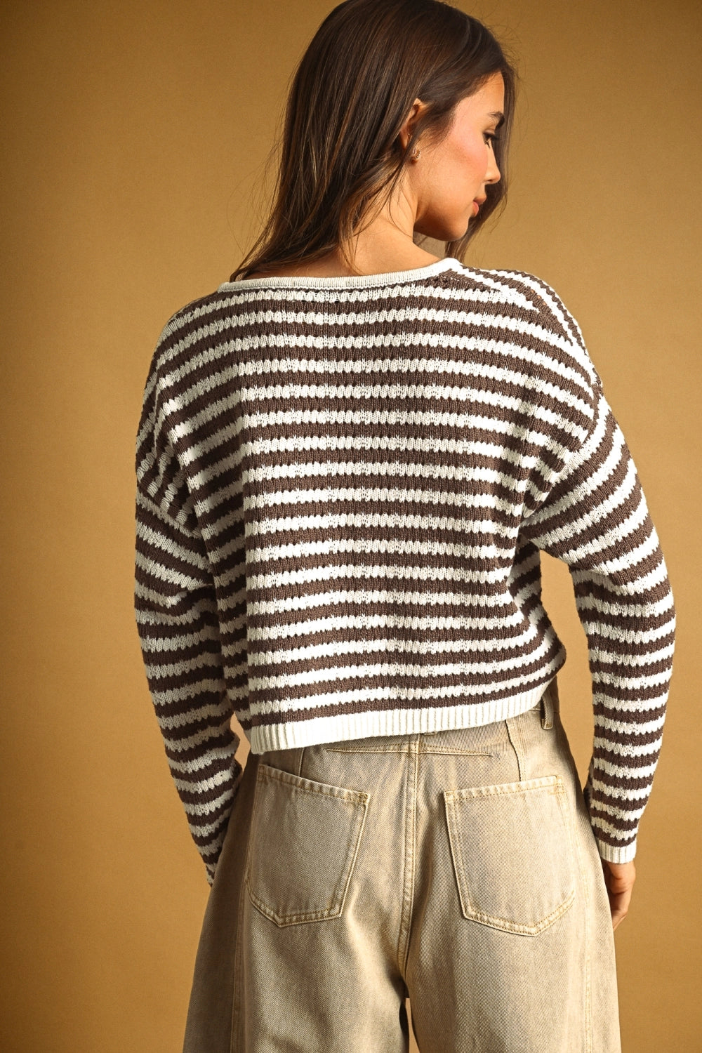 A woman shown from the back wearing a brown and white horizontally striped knit sweater and light beige jeans, set against a solid warm tan background.