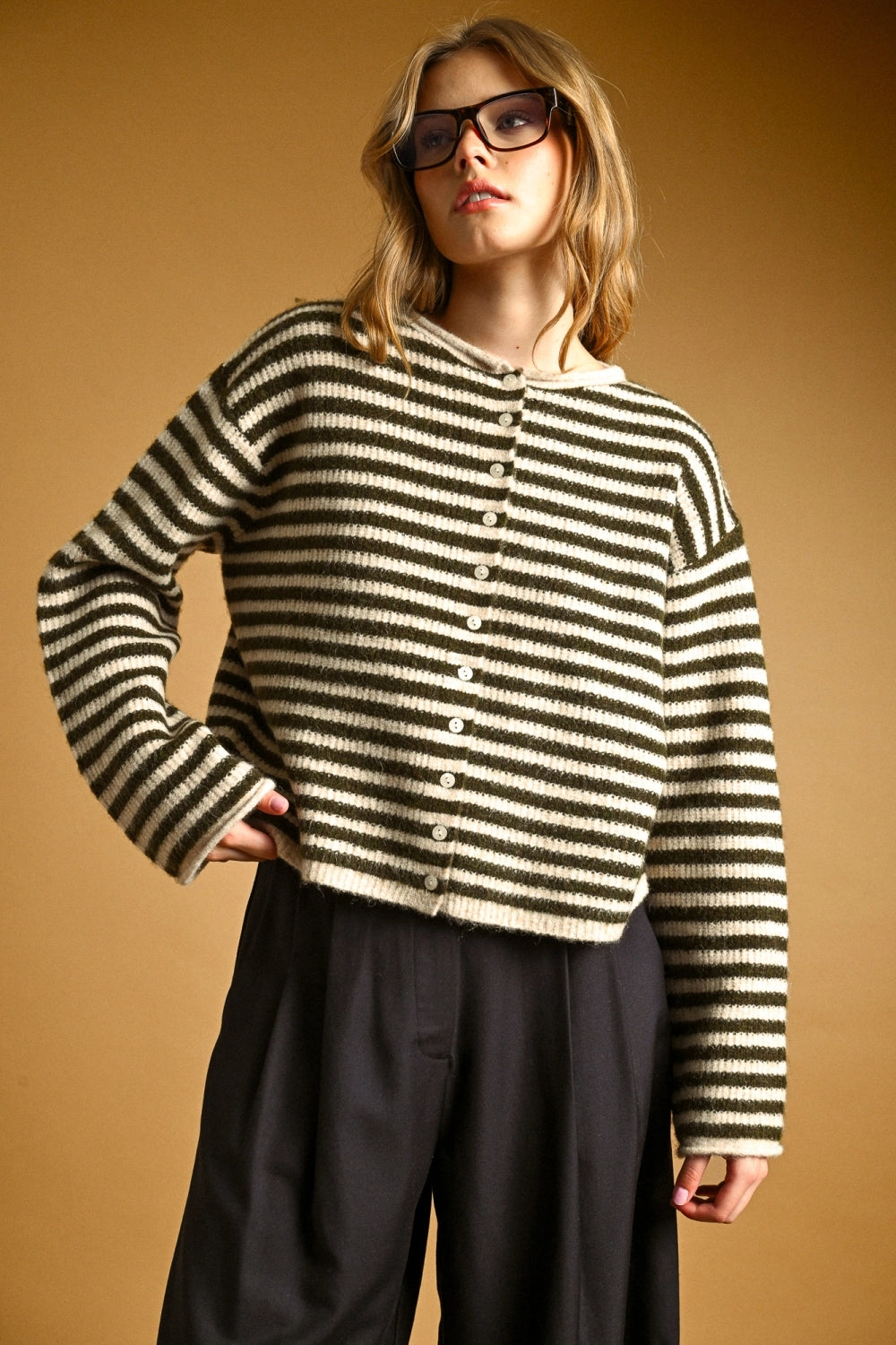 A woman wearing thick black-framed glasses and an oversized black and cream striped knit button-front top, posed against a warm tan studio background.