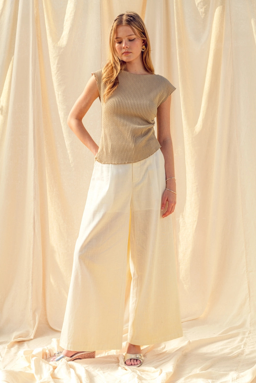 Woman wearing a beige top and white pants standing against a beige curtain.