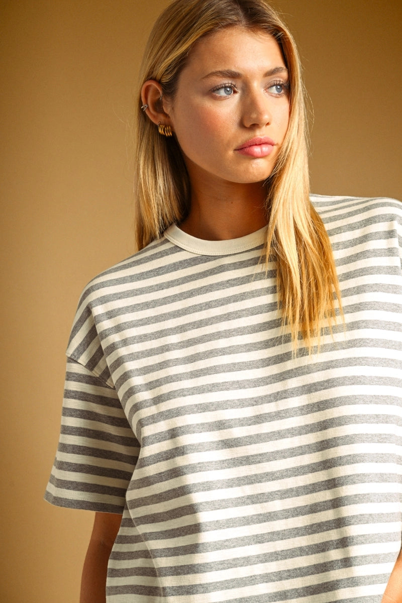 A woman wearing an oversized short-sleeve striped t-shirt with horizontal gray and off-white stripes, set against a solid warm tan background.