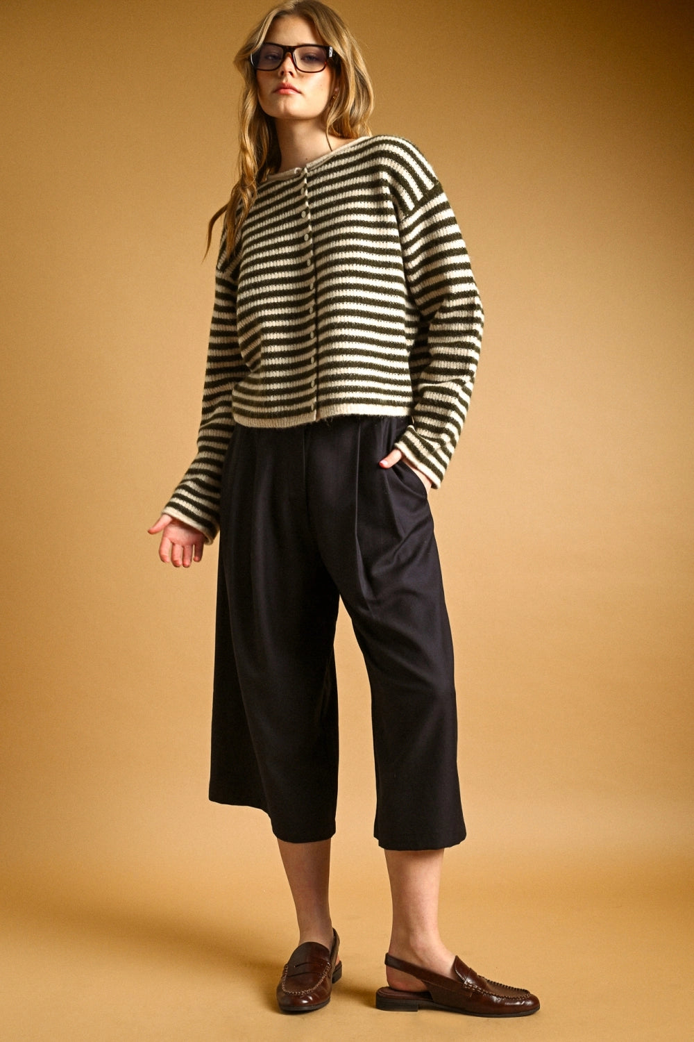 A woman wearing thick black-framed glasses and an oversized black and cream striped knit button-front top, posed against a warm tan studio background.