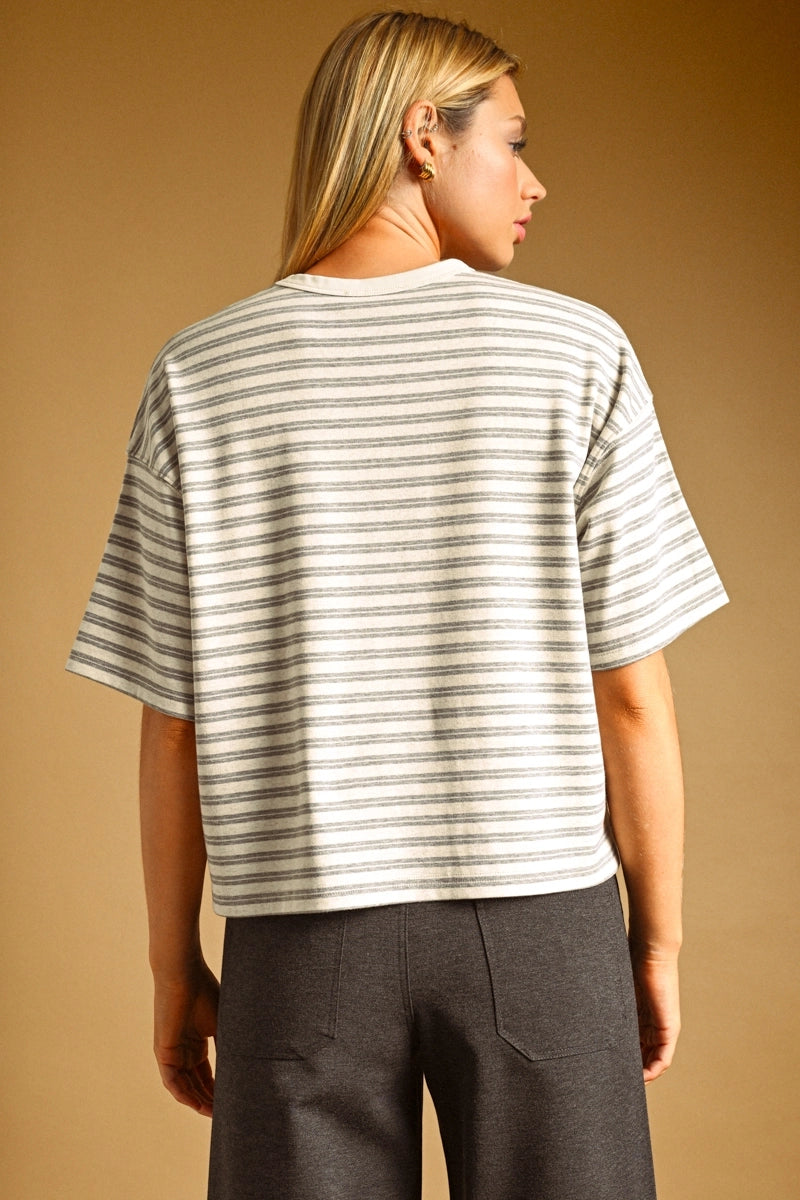 A woman shown from the back wearing a short-sleeve striped knit top with dark gray pants, set against a solid warm tan studio background.