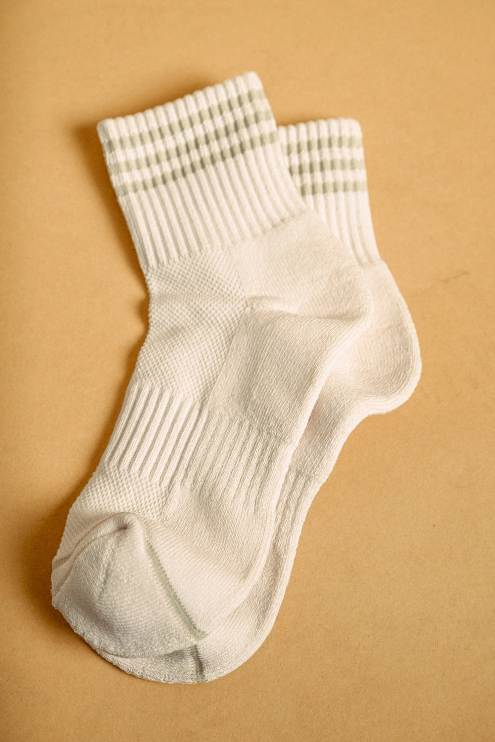A pair of off-white crew socks with sage stripes near the cuff, laid flat on a warm tan background.