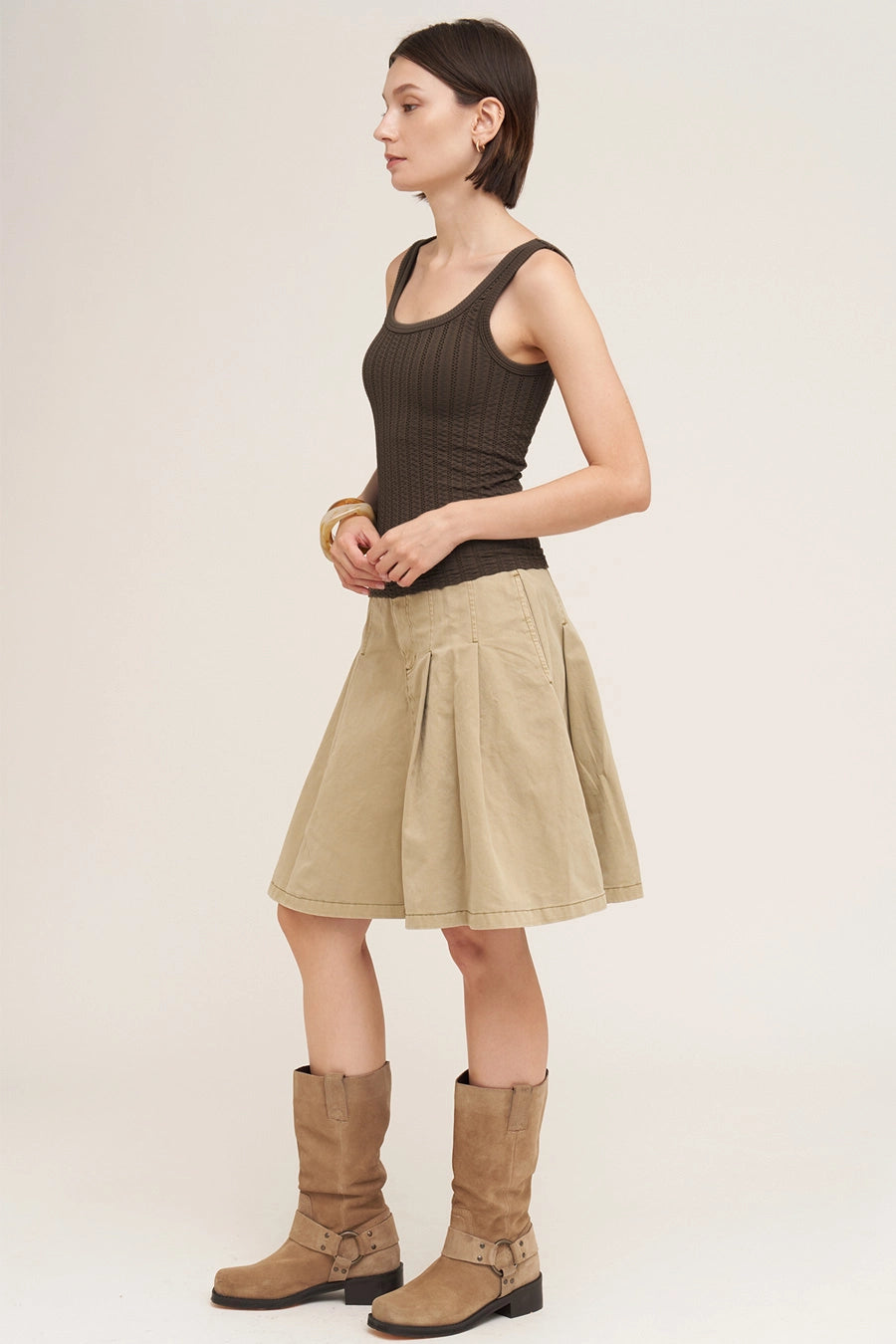 A woman stands in profile wearing a dark brown ribbed tank top, a beige pleated A-line skirt, and tan knee-high boots against a plain light beige studio background.