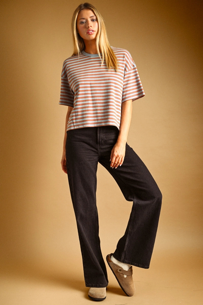 A woman modeling a short-sleeve crewneck striped t-shirt in muted pink, blue, and white, paired with black wide-leg jeans and beige slip-on shoes, against a warm brown studio backdrop.