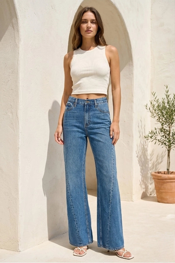 Woman wearing a white top and blue jeans standing in a sunlit outdoor setting.