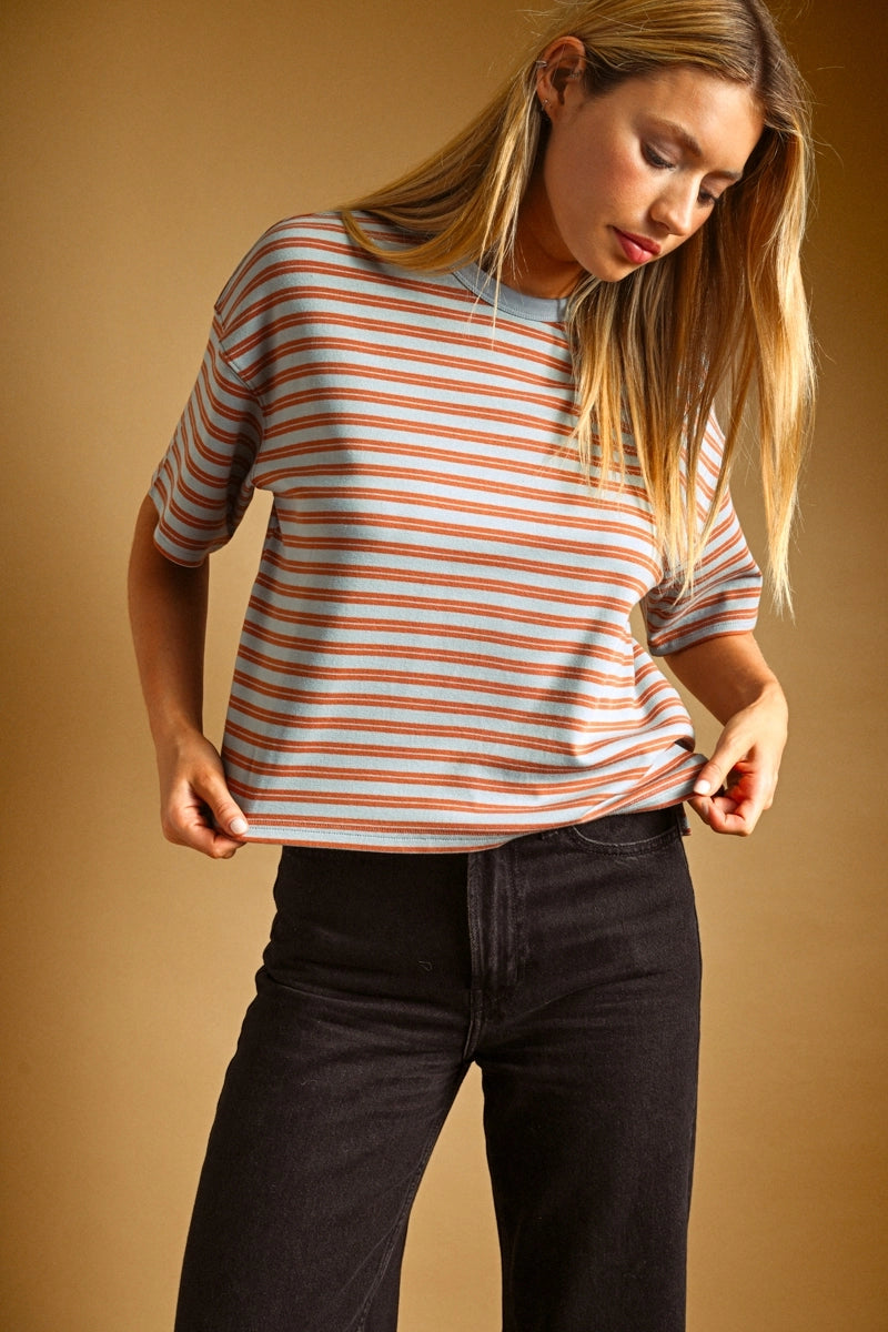 A woman wearing a short-sleeve crewneck t-shirt with thin orange and white horizontal stripes, paired with black jeans, set against a solid warm brown studio background.