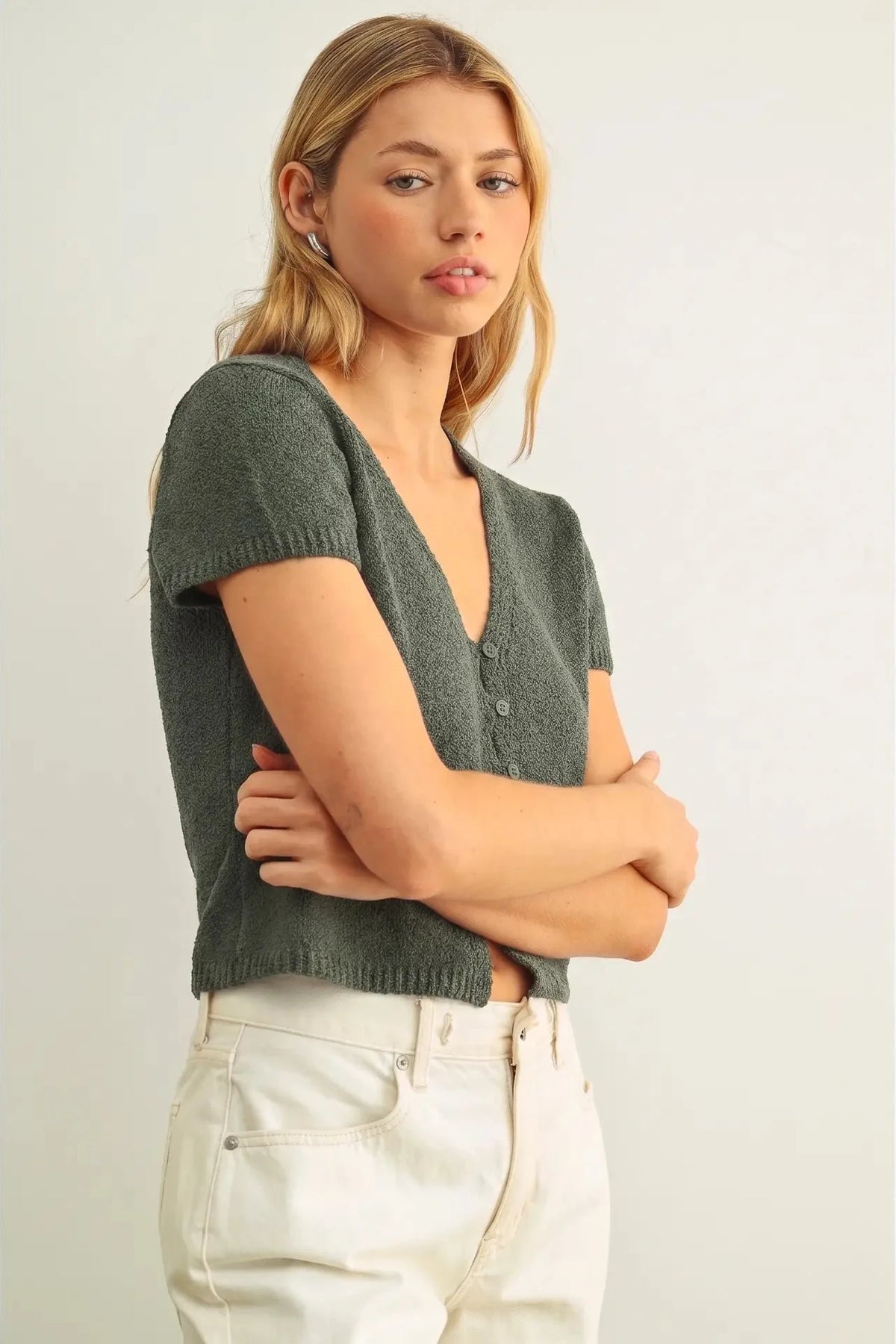 Woman wearing a green knitted top and white pants against a plain background