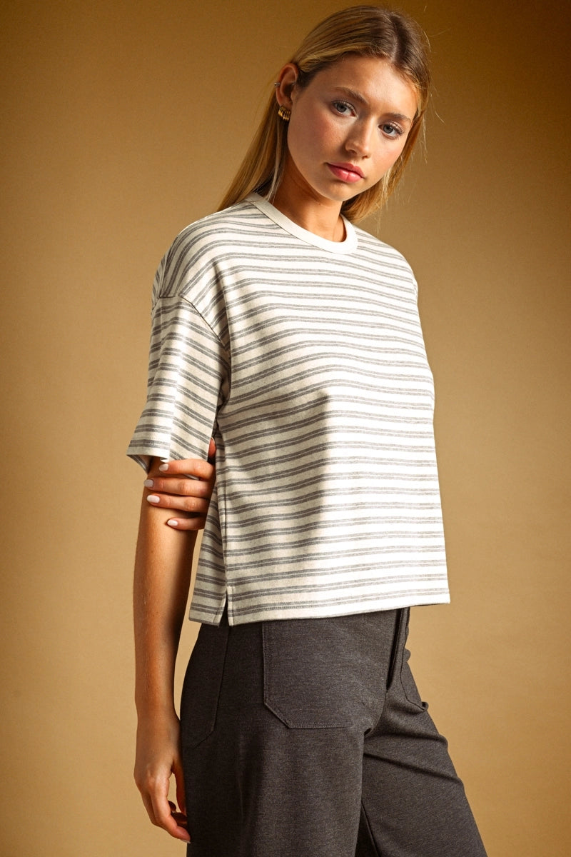 A woman modeling a short-sleeve crewneck top with thin horizontal gray stripes on an off-white base, paired with dark gray pants, against a solid warm brown studio background.