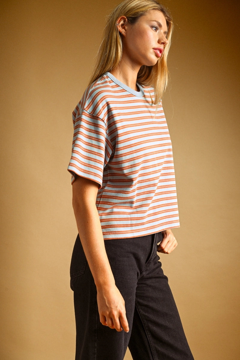 A woman wearing a short-sleeve crewneck t-shirt with thin orange and white horizontal stripes, paired with black jeans, set against a solid warm brown studio background.