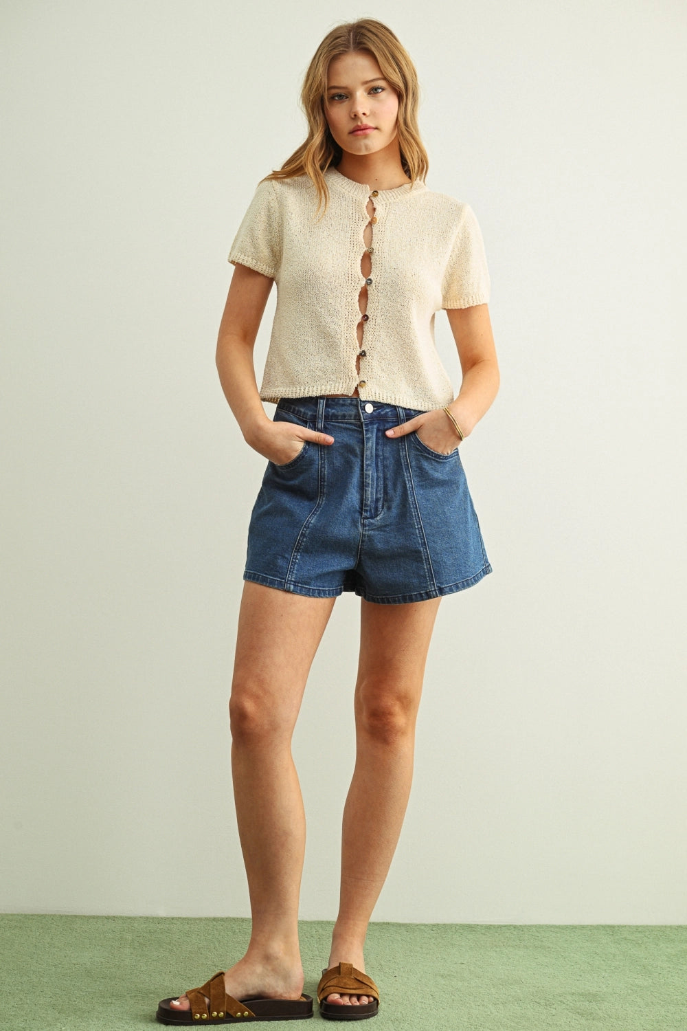 Woman wearing a beige button-up shirt and blue denim shorts on a light green background
