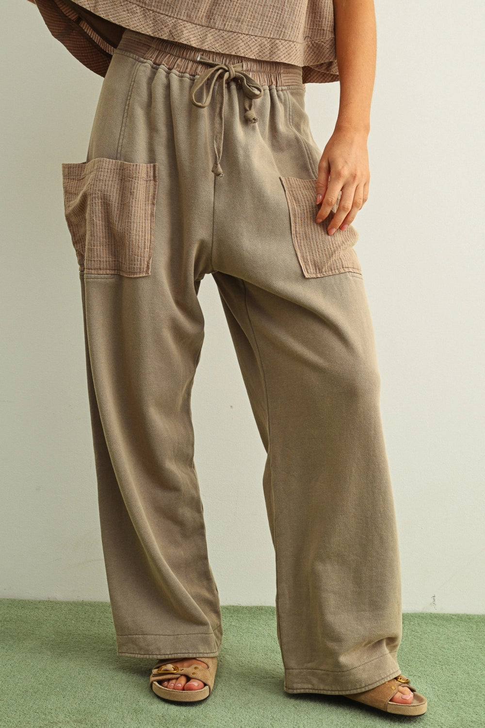 A person wearing loose, wide-leg taupe pants with a drawstring waist and large patch pockets, paired with a matching top and sandals, against a plain light wall and green floor.