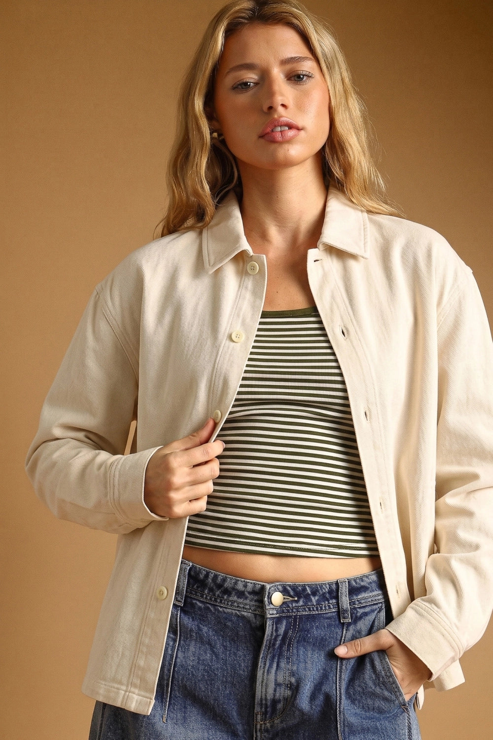 A woman wearing a light beige button-up overshirt over a green and white striped crop top, paired with blue jeans, set against a solid warm brown studio background.