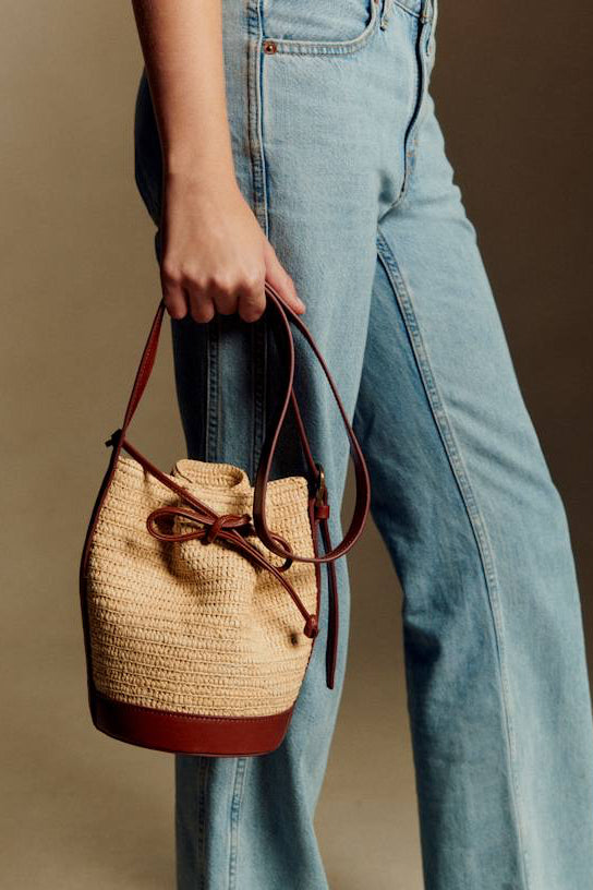 Person holding a straw bucket bag with brown leather accents, wearing blue jeans.