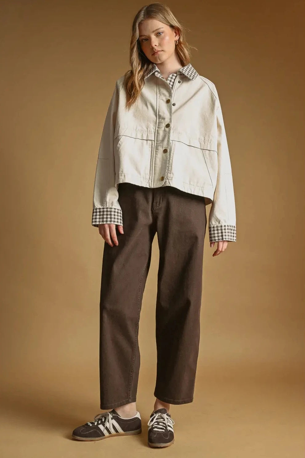 Woman wearing a light-colored jacket with plaid accents and brown pants on a beige background