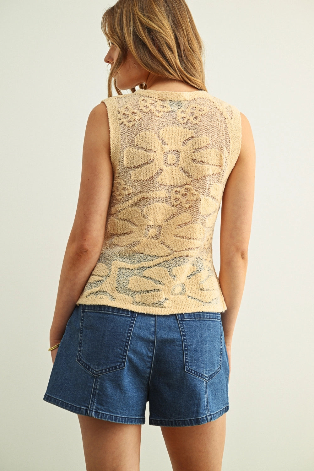 Woman wearing a beige sleeveless top with floral pattern and denim shorts on a plain background